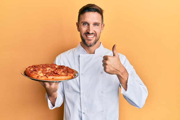 Hombre guapo con barba cocinero profesional sosteniendo pizza italiana sonriendo feliz y positivo, pulgar hacia arriba haciendo excelente y signo de aprobación  - Foto, imagen