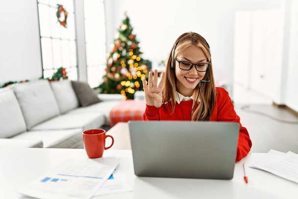 若いですcaucasian女の子に座っていますテーブルの上で作業ノートパソコンを使用してクリスマスツリーによって表示され、指の数4で指摘しながら、自信と幸せ.  - 写真・画像