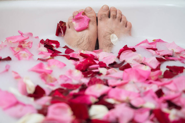 Funny picture of a man taking a relaxing bath. Close-up of male feet in a bath with foam and rose petals - Fotó, kép