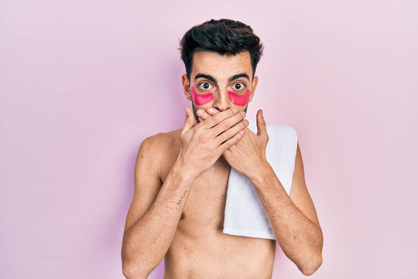 Jonge Spaanse man shirtloos dragen handdoek en oogzakken pleisters geschokt bedekken mond met handen voor fout. geheim concept.  - Foto, afbeelding