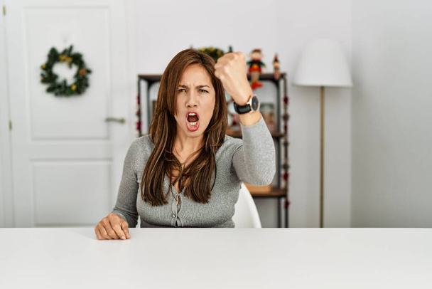 Νεαρή Λατίνα γυναίκα κάθεται στο τραπέζι με χριστουγεννιάτικη διακόσμηση θυμωμένος και τρελός ανύψωση γροθιά απογοητευμένοι και έξαλλος, ενώ φωνάζει με θυμό. οργή και επιθετική αντίληψη.  - Φωτογραφία, εικόνα
