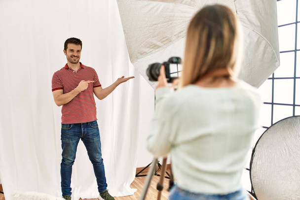 Mulher fotógrafa falando fotos de homem posando como modelo no estúdio de fotografia espantado e sorrindo para a câmera ao apresentar com a mão e apontando com o dedo.  - Foto, Imagem