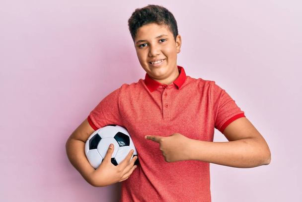 Teenager hispánský chlapec drží fotbalový míč s úsměvem šťastný ukazující rukou a prstem  - Fotografie, Obrázek
