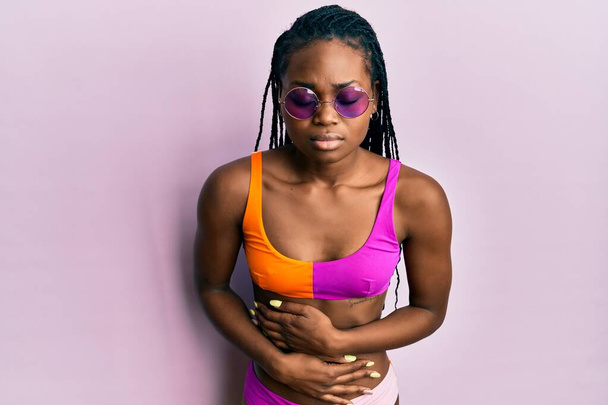 Jonge Afro-Amerikaanse vrouw draagt bikini en zonnebril met de hand op de maag omdat indigestie, pijnlijke ziekte zich niet goed voelt. pijnconcept.  - Foto, afbeelding