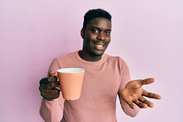 Hombre negro guapo bebiendo una taza de café celebrando el logro con sonrisa feliz y la expresión ganadora con la mano levantada  - Foto, imagen