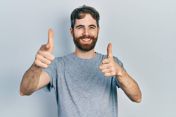 Кавказький чоловік з бородою у звичайній сірій сорочці вказує пальцями на камеру з щасливим і кумедним обличчям. добра енергія й трубопроводи.  - Фото, зображення