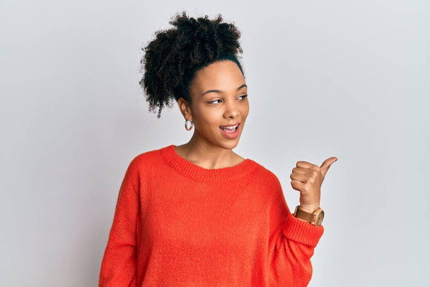 Jeune fille afro-américaine portant des vêtements décontractés souriant avec le visage heureux regardant et pointant vers le côté avec le pouce vers le haut.  - Photo, image