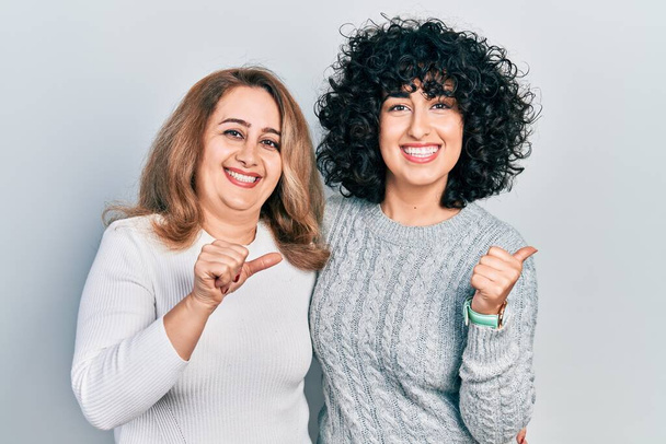 Mutter und Tochter im Nahen Osten in lässiger Kleidung, die nach hinten zeigt, mit erhobener Hand und Daumen, selbstbewusst lächelnd  - Foto, Bild