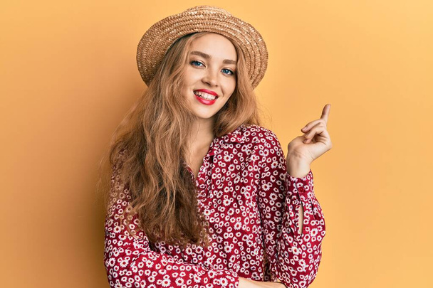 Beautiful blonde caucasian woman wearing summer hat with a big smile on face, pointing with hand and finger to the side looking at the camera.  - Foto, Imagen