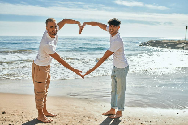 Young gay couple smiling happy doing heart symbol with arms at the beach. - Photo, Image