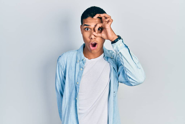 Young african american guy wearing casual clothes doing ok gesture shocked with surprised face, eye looking through fingers. unbelieving expression.  - Photo, Image