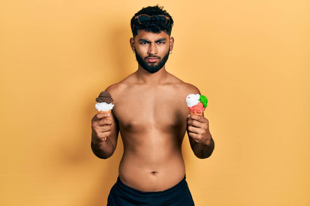Mayo giyen sakallı bir Arap şüpheci ve gergin iki dondurma külahı yiyor, sorun yüzünden surat asıyor. negatif kişi.  - Fotoğraf, Görsel