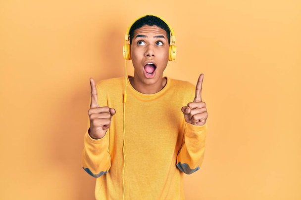 Jonge Afro-Amerikaanse man luisteren naar muziek met behulp van een koptelefoon verbaasd en verrast omhoog te kijken en te wijzen met de vingers en opgeheven armen.  - Foto, afbeelding