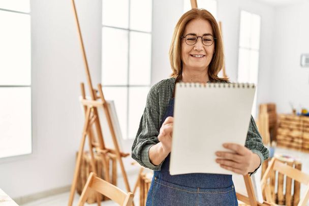 Middelbare leeftijd kunstenaar vrouw glimlachen gelukkig schilderij op notebook in kunststudio. - Foto, afbeelding
