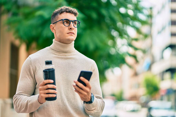 Młody Hiszpan nosi okulary za pomocą smartfona i trzyma butelkę wody w mieście. - Zdjęcie, obraz