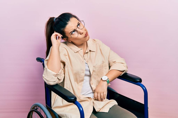 Young hispanic woman sitting on wheelchair confuse and wondering about question. uncertain with doubt, thinking with hand on head. pensive concept.  - Photo, image