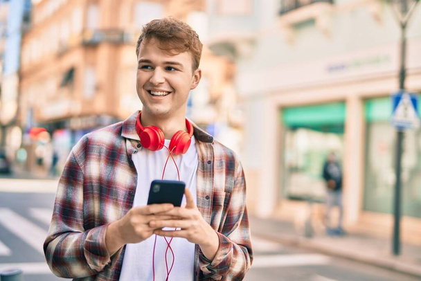 Jeune homme caucasien souriant heureux en utilisant un smartphone à la ville. - Photo, image