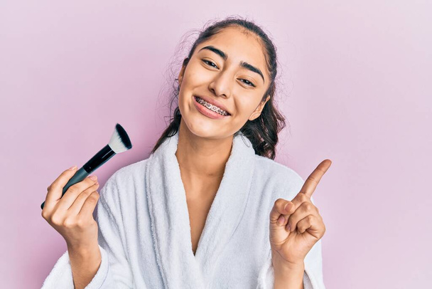 Латиноамериканская девочка-подросток с зубными брекетами в халате, держа кисть для макияжа, улыбаясь счастливо указывая рукой и пальцем в сторону  - Фото, изображение