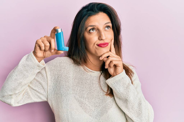 Beautiful hispanic woman holding medical asthma inhaler serious face thinking about question with hand on chin, thoughtful about confusing idea  - Photo, Image