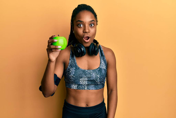Afrikkalainen amerikkalainen nainen punottu hiukset yllään urheiluvaatteet ja kuulokkeet syö vihreää omenaa peloissaan ja hämmästynyt suu auki yllätys, epäusko kasvot  - Valokuva, kuva