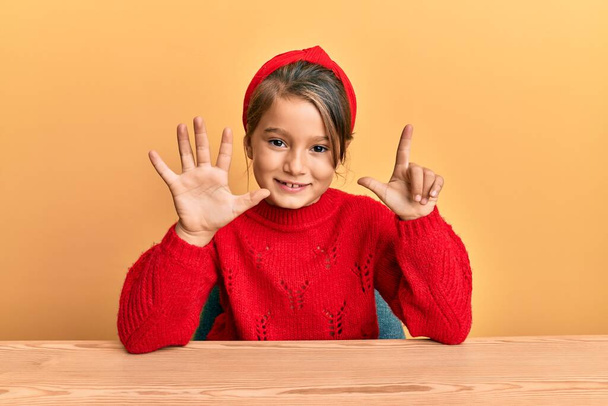 Μικρό όμορφο κορίτσι φορώντας casual ρούχα κάθεται στο τραπέζι δείχνει και δείχνοντας προς τα πάνω με τα δάχτυλα αριθμό επτά, ενώ χαμογελά αυτοπεποίθηση και χαρούμενος.  - Φωτογραφία, εικόνα