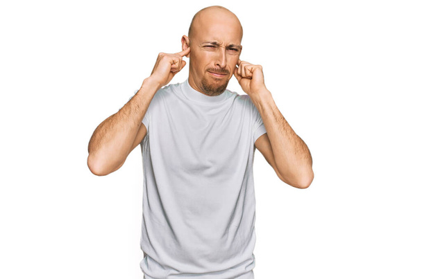 Hombre calvo con barba vistiendo camiseta blanca casual cubriendo las orejas con los dedos con expresión molesta por el ruido de la música alta. concepto de sordo.  - Foto, imagen