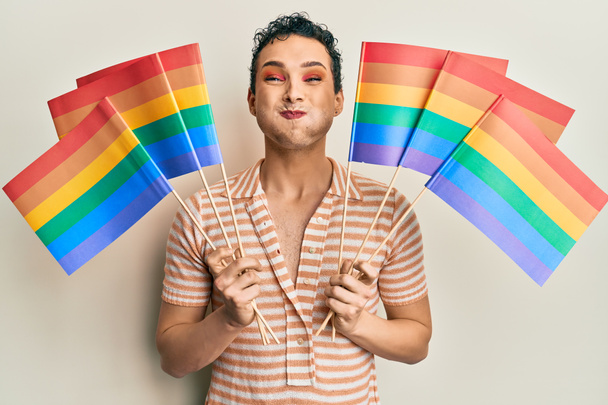 Hombre guapo usando maquillaje sosteniendo banderas de arco iris lgbtq soplando mejillas con cara divertida. boca inflada con aire, captación de aire.  - Foto, imagen