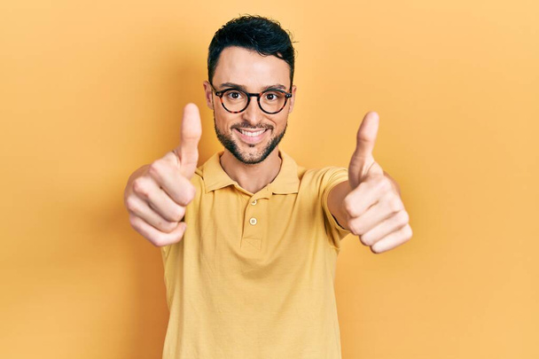 Jonge Spaanse man met casual kleding en bril die positief gebaar met de hand goedkeurt, glimlacht lachend en blij voor succes. winnaar gebaar.  - Foto, afbeelding