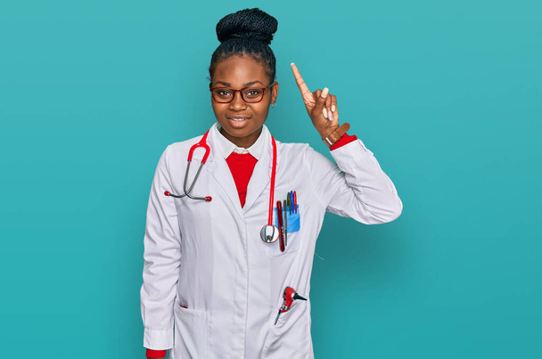 Молодая африканская американка в форме врача и стетоскопе показывает и показывает пальцем номер один, улыбаясь уверенно и счастливо.  - Фото, изображение