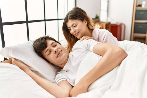 Νεαρή καυκάσια γυναίκα χαμογελά χαρούμενη ξυπνώντας το αγόρι της ξαπλωμένο στο κρεβάτι στο σπίτι. - Φωτογραφία, εικόνα