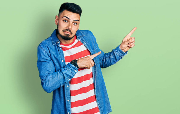 Junger hispanischer Mann mit Bart in lässiger Jeansjacke, der besorgt und nervös mit beiden Händen zur Seite zeigt, besorgter und überraschter Ausdruck  - Foto, Bild