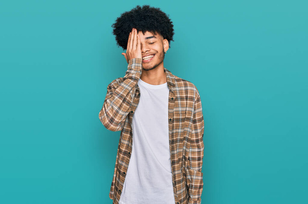 Jonge Afro-Amerikaanse man met afro haar in casual kleding die een oog bedekt met de hand, zelfverzekerde glimlach op het gezicht en verrassing emotie.  - Foto, afbeelding