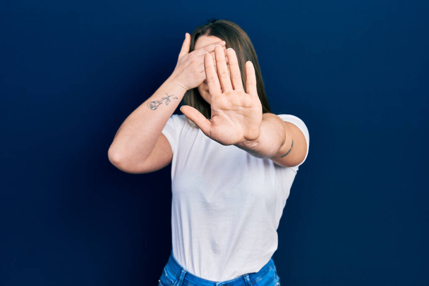 Молодая латиноамериканка в обычной белой футболке, закрывающей глаза руками и делающей остановку жеста с грустным и испуганным выражением лица. смущенная и негативная концепция.  - Фото, изображение