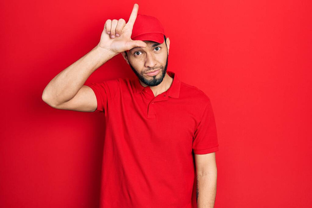 Hispanic man with beard wearing delivery uniform and cap making fun of people with fingers on forehead doing loser gesture mocking and insulting.  - Photo, Image