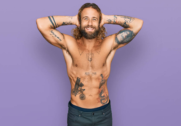 Hombre guapo con barba y pelo largo de pie sin camisa mostrando tatuajes relajante y estiramiento, brazos y manos detrás de la cabeza y el cuello sonriendo feliz  - Foto, imagen