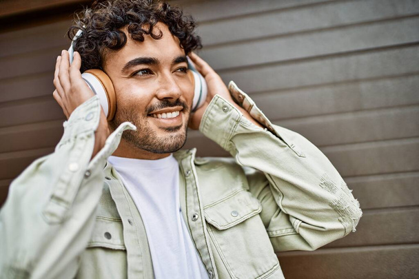 Schöner hispanischer Mann mit Bart, der an einem sonnigen Tag im Freien glücklich lächelt und Kopfhörer trägt - Foto, Bild