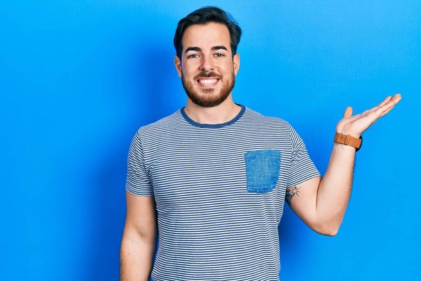 bel homme caucasien avec barbe portant casual t-shirt rayé souriant gai présentation et pointant avec la paume de la main regardant la caméra.  - Photo, image