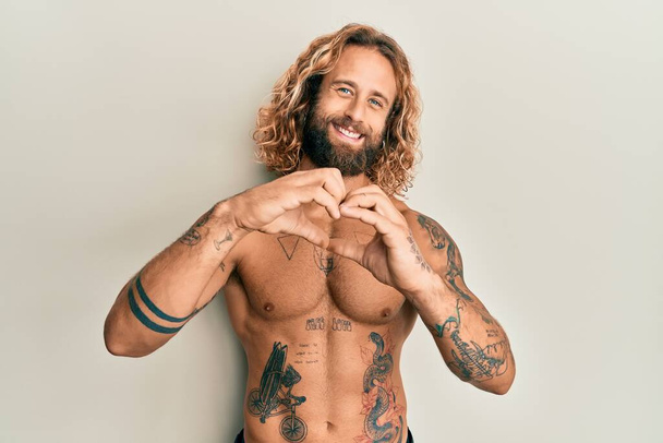 Jóképű férfi szakállal és hosszú hajjal, félmeztelen, tetovált, szerelmes tetoválásokkal, szív szimbólum alakú kezekkel. romantikus koncepció.  - Fotó, kép