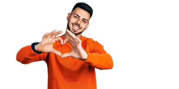 ひげを生やした若いヒスパニック系の男性は、手でハートシンボルの形をして愛に笑みを浮かべてカジュアルなオレンジのセーターを着ています。ロマンチックなコンセプト.  - 写真・画像