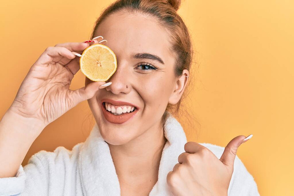 Mujer rubia joven con albornoz sosteniendo limón sobre el ojo apuntando el pulgar hacia el lado sonriendo feliz con la boca abierta  - Foto, Imagen