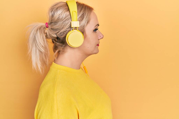 Młoda blondynka słucha muzyki za pomocą słuchawek patrząc na bok, relaks profil pozować z naturalną twarz z pewnym uśmiechem.  - Zdjęcie, obraz