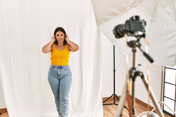 Young beautiful hispanic woman posing as model at photography studio covering ears with fingers with annoyed expression for the noise of loud music. deaf concept.  - Zdjęcie, obraz
