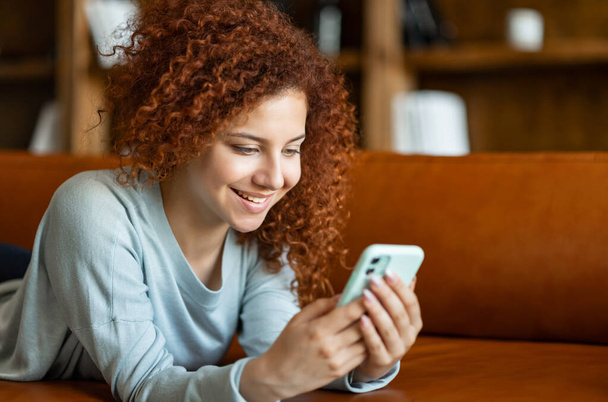 Счастливая молодая привлекательная кудрявая рыжая женщина с мобильного телефона - Фото, изображение