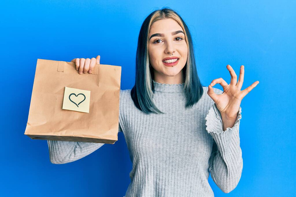 Fiatal modern lány gazdaság elvenni papírzacskó szívjegyzet csinál rendben jel ujjakkal, mosolygós barátságos gesztus kiváló szimbólum  - Fotó, kép