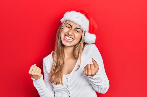 クリスマスの帽子をかぶって美しいヒスパニック系の女性は非常に満足し、腕を上げて勝者のジェスチャーを行う興奮し、笑顔と成功のために叫んでいます。お祝いのコンセプト.  - 写真・画像