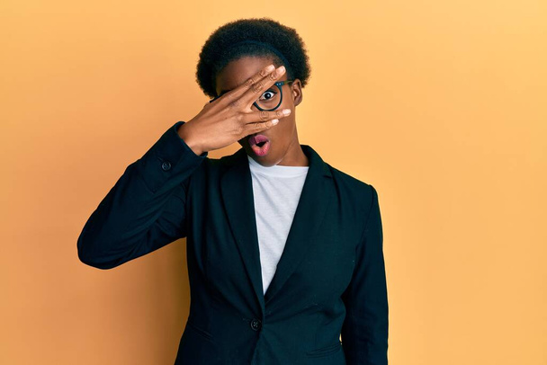 Jovem afro-americana vestindo jaqueta de negócios e óculos espreitando em choque cobrindo rosto e olhos com a mão, olhando através dos dedos com medo  - Foto, Imagem