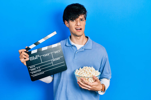 Knappe hippe jongeman die popcorn eet met een klapbord vol bioscoopgeluiden. twijfelconcept.  - Foto, afbeelding