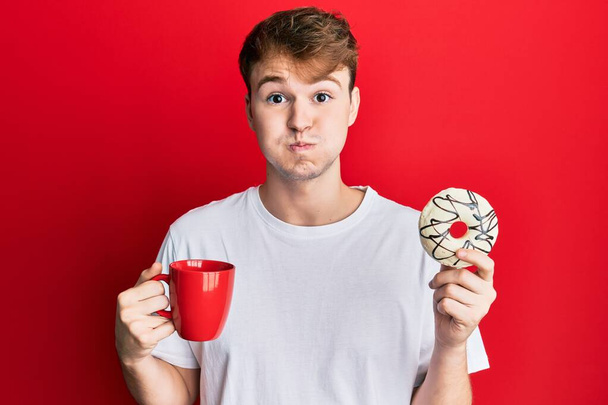 Young caucasian man drinking a cup of coffee and croissant puffing cheeks with funny face. mouth inflated with air, catching air.  - Photo, Image