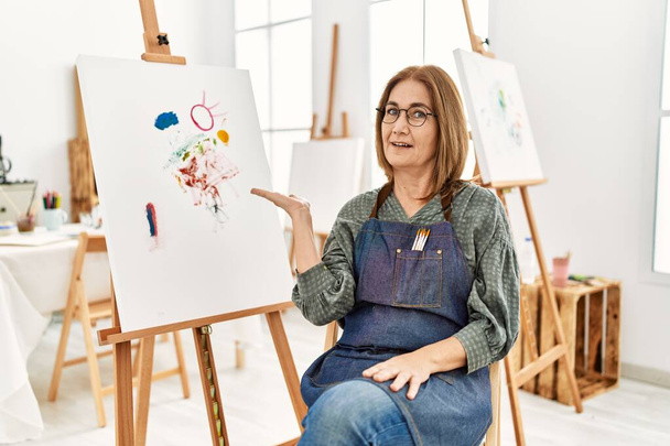 Künstlerin mittleren Alters im Atelier lächelt fröhlich und zeigt mit der Handfläche in die Kamera.  - Foto, Bild