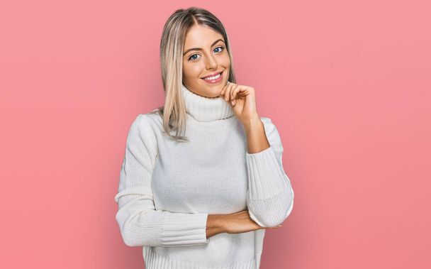 Beautiful blonde woman wearing casual turtleneck sweater looking confident at the camera with smile with crossed arms and hand raised on chin. thinking positive.  - Photo, Image